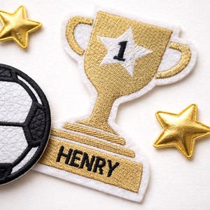 May include: An embroidered patch shaped like a gold trophy with a white star and the number 1, featuring the name HENRY in black text on the base. Beside it is a black and white soccer ball patch and two small gold star patches.