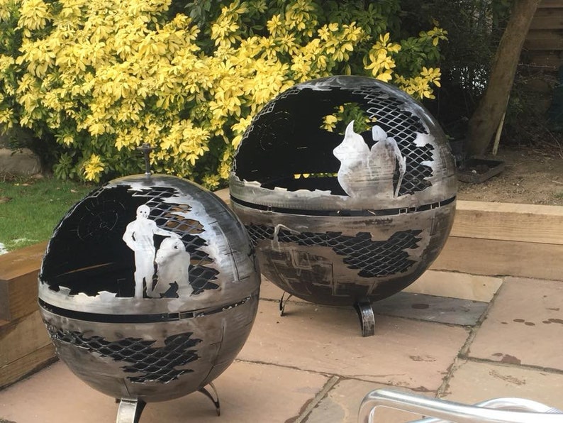 May include: Two metal fire pit spheres with intricate cutouts. One sphere features a Star Wars scene with Luke Skywalker and a character from the movie. The other sphere has a more abstract design.