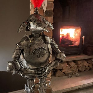 Op de afbeelding: Een zilveren metalen ridderfiguur met een harnas en een zwaard. De ridder heeft een rode veer op zijn helm en staat voor een open haard.