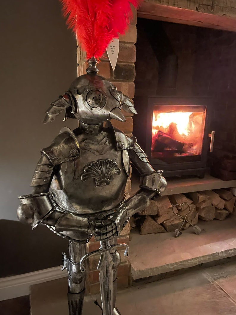 Op de afbeelding: Een zilveren metalen ridderfiguur met een harnas en een zwaard. De ridder heeft een rode veer op zijn helm en staat voor een open haard.