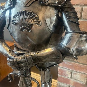 Op de afbeelding: Een metalen sculptuur van een ridder in harnas, die een zwaard vasthoudt. De ridder draagt een helm en een borstplaat met een schelpvormig ontwerp. Het beeld staat op een houten voet.