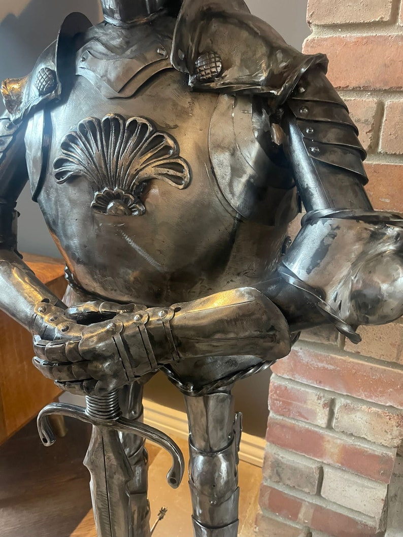 Op de afbeelding: Een metalen sculptuur van een ridder in harnas, die een zwaard vasthoudt. De ridder draagt een helm en een borstplaat met een schelpvormig ontwerp. Het beeld staat op een houten voet.