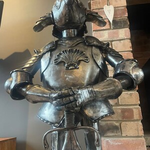 Op de afbeelding: Een metalen ridderfiguur met een harnas met een rode veer op zijn helm. De ridder houdt een zwaard vast en heeft een schelpontwerp op zijn borst.