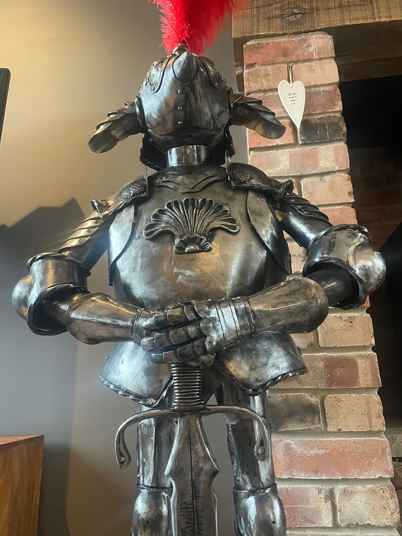 Op de afbeelding: Een metalen ridderfiguur met een harnas met een rode veer op zijn helm. De ridder houdt een zwaard vast en heeft een schelpontwerp op zijn borst.
