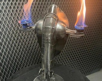 May include: A metal sculpture of a spacecraft with two flames. The sculpture is silver and has a circular base with a symbol. The flames are blue and orange. The background is a metal mesh.