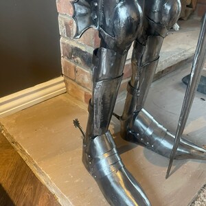 Op de afbeelding: Close-up van de benen van een metalen ridderstandbeeld, met gedetailleerde bepantsering met een puntige teen en een sporen aan &eacute;&eacute;n voet.