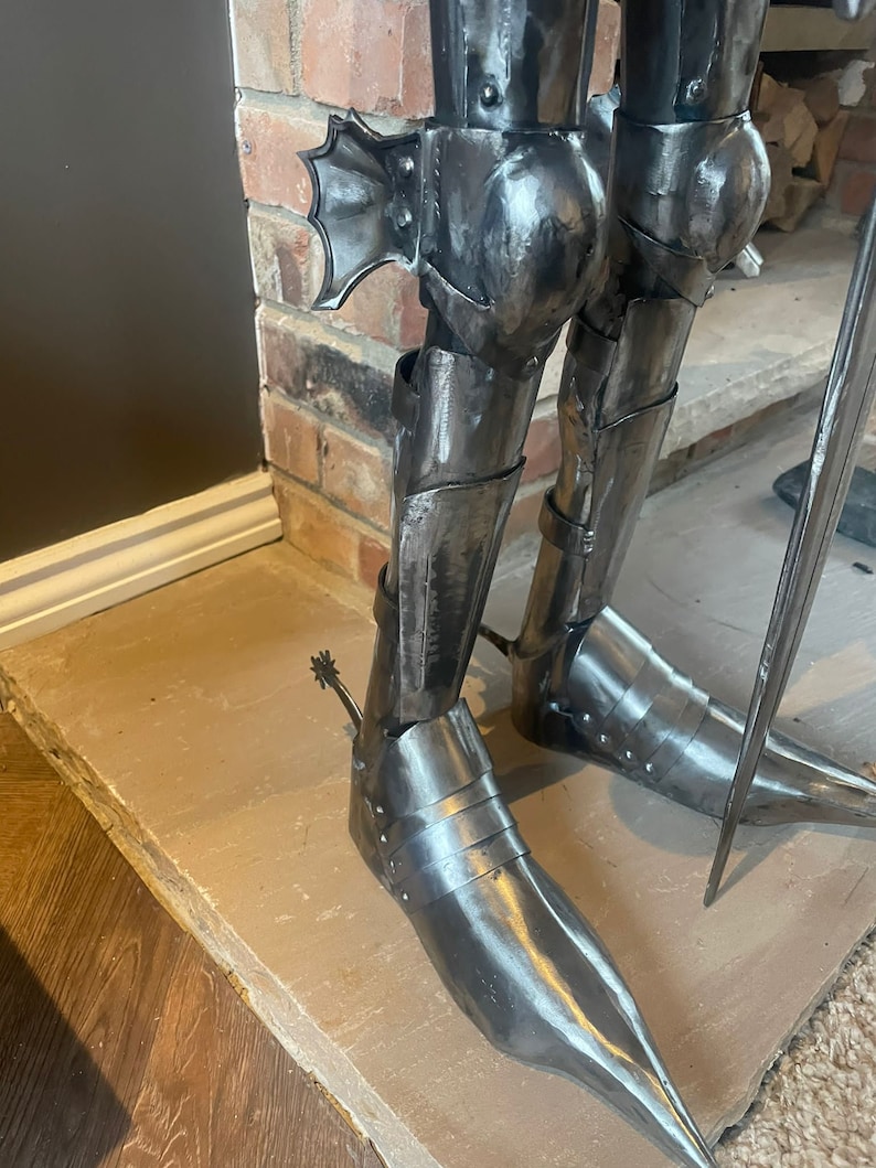 Op de afbeelding: Close-up van de benen van een metalen ridderstandbeeld, met gedetailleerde bepantsering met een puntige teen en een sporen aan &eacute;&eacute;n voet.