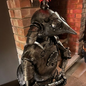 Op de afbeelding: Een metalen sculptuur van een ridder in harnas, met een rode verenpluim op zijn helm. De ridder houdt een zwaard in zijn rechterhand en een schild aan zijn linkerarm. Het beeld is gemaakt van gerecycled metaal en heeft een rustieke, verweerde uitstraling.