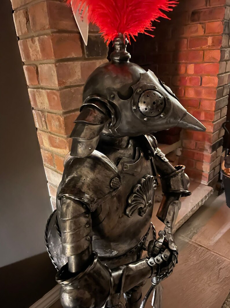 Op de afbeelding: Een metalen sculptuur van een ridder in harnas, met een rode verenpluim op zijn helm. De ridder houdt een zwaard in zijn rechterhand en een schild aan zijn linkerarm. Het beeld is gemaakt van gerecycled metaal en heeft een rustieke, verweerde uitstraling.