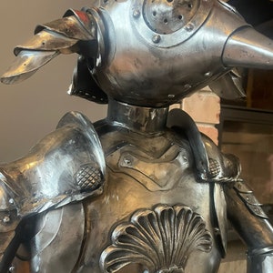Op de afbeelding: Een metalen sculptuur van een ridder die een harnas draagt met een helm in de vorm van een vogelkop. Het harnas is versierd met een schelpontwerp op de borst.