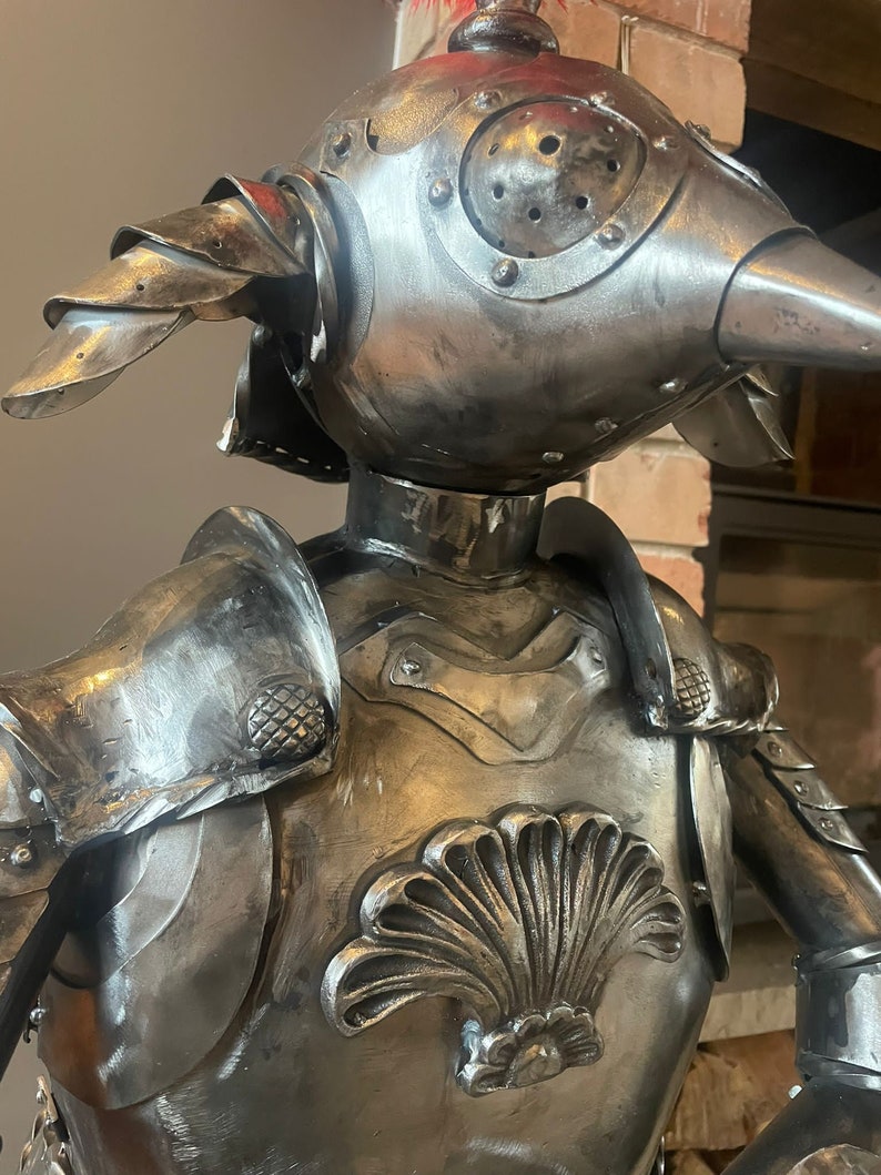 Op de afbeelding: Een metalen sculptuur van een ridder die een harnas draagt met een helm in de vorm van een vogelkop. Het harnas is versierd met een schelpontwerp op de borst.