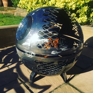 May include: A metal fire pit shaped like the Death Star from Star Wars. The fire pit has a mesh design and is burning with flames.
