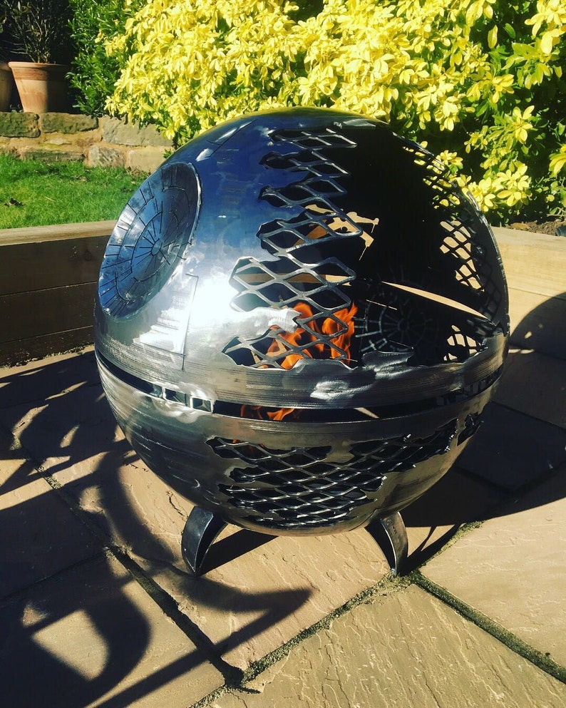 May include: A metal fire pit shaped like the Death Star from Star Wars. The fire pit has a mesh design and is burning with flames.