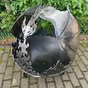 Puede incluir: Un brasero de metal negro con forma de dragón con las alas extendidas. La cabeza del dragón está mirando hacia adelante y su boca está abierta. El brasero está sentado en un patio de adoquines.