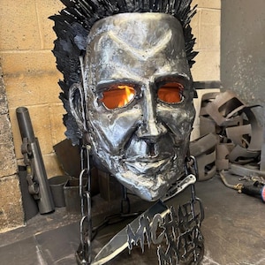 May include: A metal sculpture of a face with a black spiky hair design and orange glowing eyes. A knife is attached to the sculpture with the name "MICHAEL" and the word "HALLOWEEN" on the base.