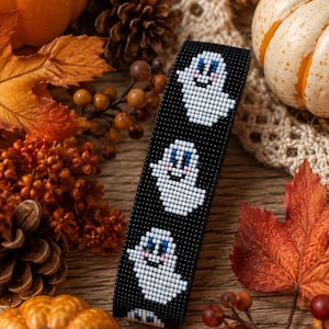 Könnte beinhalten: Ein schwarzes Perlenarmband mit einem sich wiederholenden Muster aus weißen Geistern mit blauen Augen und rosa Wangen. Die Geister bestehen aus kleinen, gleichmäßigen Perlen, die in einer Gitterstruktur auf schwarzem Grund angeordnet sind.
