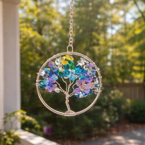 May include: A handcrafted suncatcher featuring a wire tree design within a silver-colored circular frame. The tree is adorned with colorful, faceted crystals in shades of blue, purple, yellow, and clear. It hangs from a silver chain, creating a decorative accent.