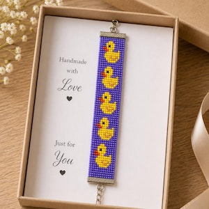 Könnte beinhalten: Ein Perlenarmband mit einem sich wiederholenden Muster aus fünf gelben Quietscheentchen mit orangefarbenen Schnäbeln auf violettem Grund. Das Armband ist auf einer weißen Karte mit dem schwarzen Text Handmade with Love und Just for You befestigt.