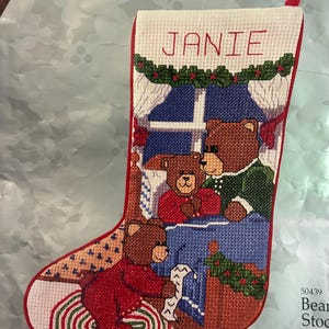 May include: Cross-stitch Christmas stocking featuring a family of teddy bears. The stocking is red with a white top, embroidered with the name "JANIE" and a festive garland. Bears are depicted in a window scene.