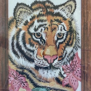 May include: A framed embroidery featuring a detailed tiger face with orange, black, and white fur, accented by two pink lily flowers at the bottom corners. The artwork is encased in a simple, dark brown wooden frame with a visible grain texture.