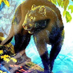 May include: A cross-stitch embroidery depicting a black panther standing on a textured surface. The panther has yellow eyes and is positioned beneath green foliage. The background features blue tones and small red and yellow floral accents.