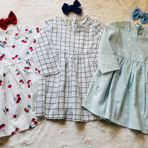 May include: Three long-sleeved dresses with matching bows. One white dress with red cherry print, one black and white grid pattern dress, and one light blue dress with white star print. The bows are red, navy, and light blue.