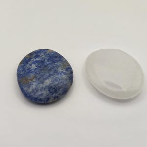 Puede incluir: Dos piedras lisas de forma ovalada. Una es de color azul oscuro con vetas blancas y grises. La otra es de un blanco lechoso translúcido. Ambas piedras están pulidas y parecen ser piedras preciosas naturales.