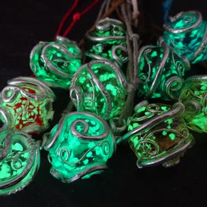 Könnte beinhalten: Eine Sammlung von leuchtend grünen und silbernen Anhängern. Jeder Anhänger hat eine kugelförmige Form mit einem wirbelnden Silberdrahtdesign. Das glasartige Material leuchtet hellgrün und erzeugt ein mystisches Aussehen. Einige Anhänger haben rote Akzente.