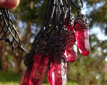 May include: A collection of red crystal pendants with black cords. Each crystal is encased in an ornate, dark metal wire wrap. The crystals vary in size and shape, with a natural, raw appearance. The background is blurred, suggesting an outdoor setting.