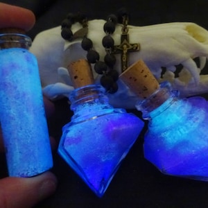 May include: Three glass bottles with cork stoppers filled with a blue and purple glittery liquid. The bottles are on a white surface with a black beaded necklace and a cross pendant.