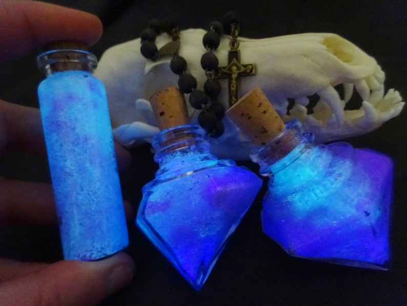 May include: Three glass bottles with cork stoppers filled with a blue and purple glittery liquid. The bottles are on a white surface with a black beaded necklace and a cross pendant.