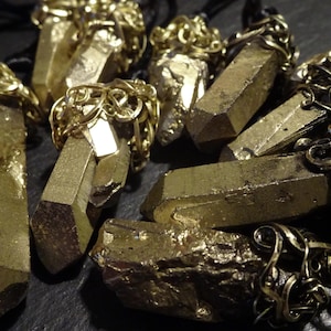 Könnte beinhalten: Eine Sammlung von goldfarbenen Kristallspitzenanhängern mit filigranen Metallakzenten. Die Anhänger sind an schwarzen Schnüren befestigt und haben verschiedene Formen und Größen.