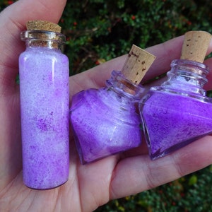 May include: Three small glass bottles with cork stoppers filled with purple and white glitter. The bottles are in a hand.