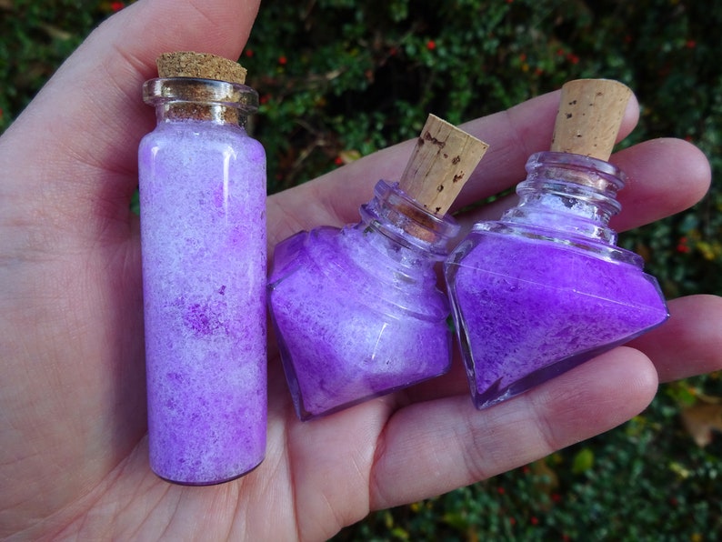 May include: Three small glass bottles with cork stoppers filled with purple and white glitter. The bottles are in a hand.