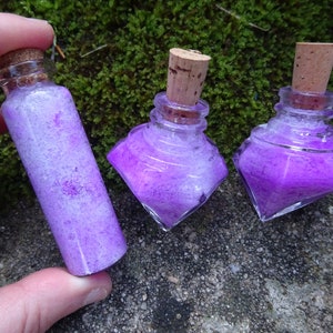 May include: Three small glass bottles with cork stoppers filled with purple glitter. The bottles are on a mossy green surface.
