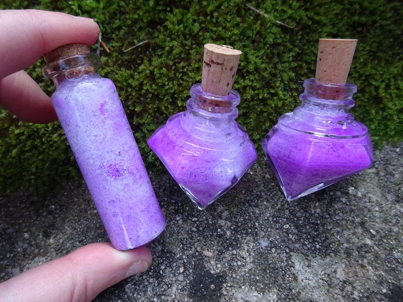 May include: Three small glass bottles with cork stoppers filled with purple glitter. The bottles are on a mossy green surface.