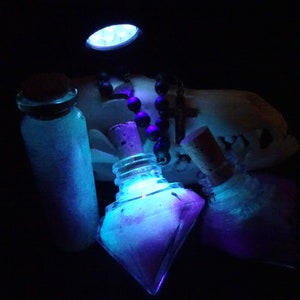 May include: Three glass bottles with cork stoppers filled with a glowing blue and purple liquid. The bottles are illuminated by a blue light source. A black beaded necklace with a cross pendant is visible in the background.