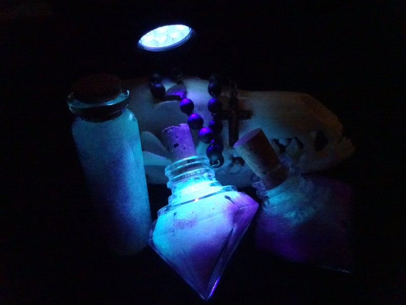 May include: Three glass bottles with cork stoppers filled with a glowing blue and purple liquid. The bottles are illuminated by a blue light source. A black beaded necklace with a cross pendant is visible in the background.