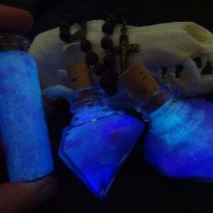 May include: Three glass bottles filled with a glowing blue liquid. The bottles have cork stoppers and are arranged on a white surface.