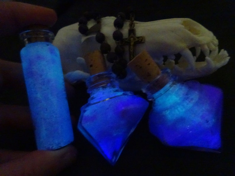 May include: Three glass bottles filled with a glowing blue liquid. The bottles have cork stoppers and are arranged on a white surface.