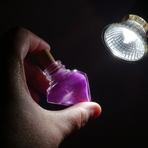 May include: A small, clear glass bottle with a cork stopper filled with purple glitter. The bottle is being held in a hand against a dark background.