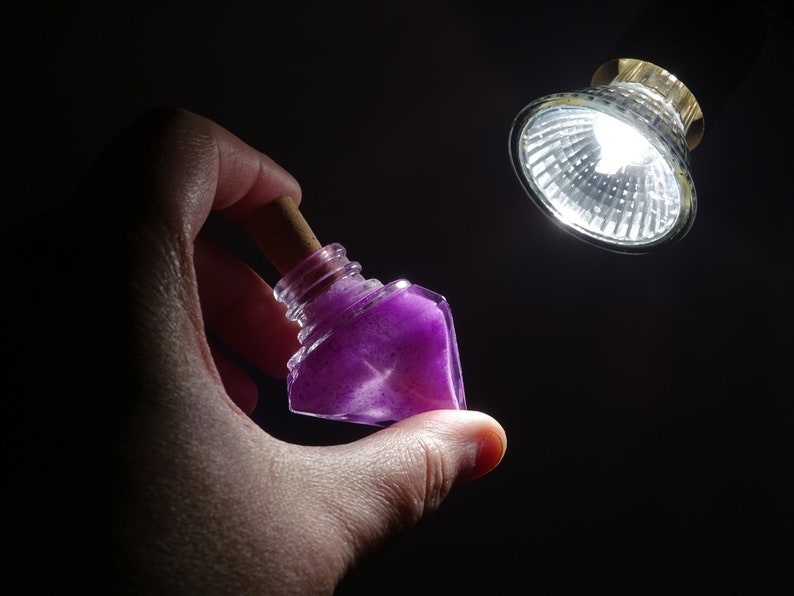 May include: A small, clear glass bottle with a cork stopper filled with purple glitter. The bottle is being held in a hand against a dark background.
