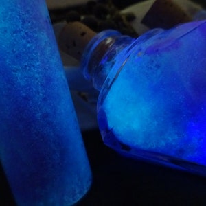 May include: Two glass bottles filled with a bright blue liquid that glows under a black light. The bottles are different shapes, one is a cylinder and the other is a diamond.