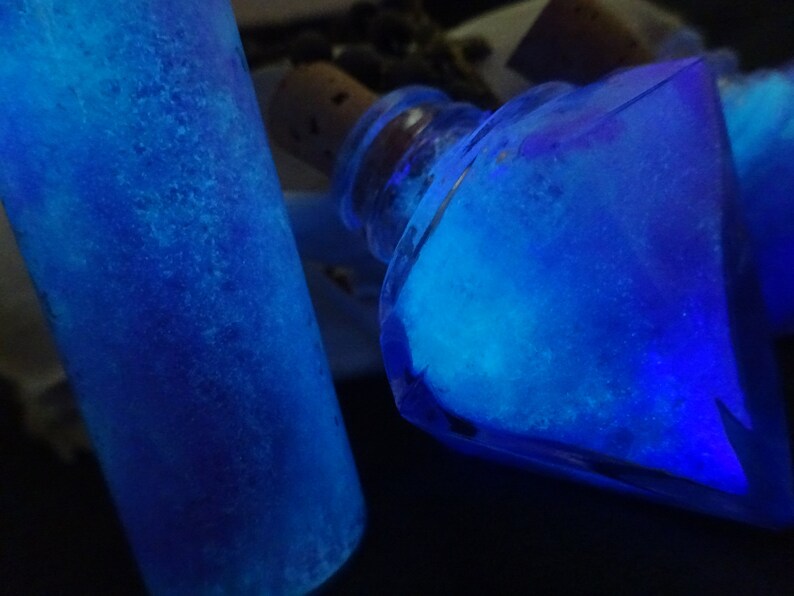 May include: Two glass bottles filled with a bright blue liquid that glows under a black light. The bottles are different shapes, one is a cylinder and the other is a diamond.