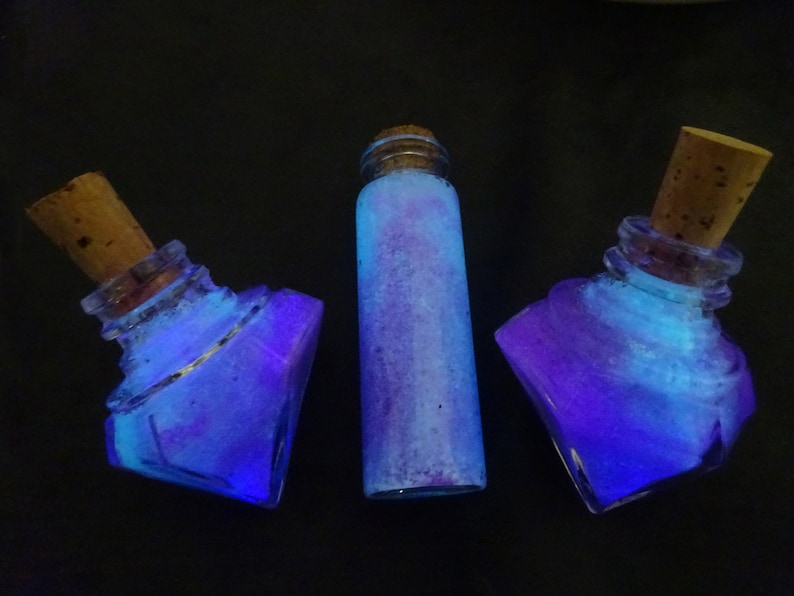 May include: Three glass bottles with cork stoppers filled with a blue and purple glittery liquid. The bottles are on a black background.