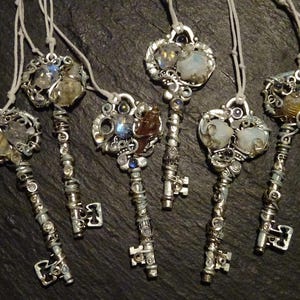 Könnte beinhalten: Sechs silberne schlüsselförmige Ornamente mit verschiedenen Kristallen und Perlen. Jeder Schlüssel hat ein anderes Design und zeigt eine Vielzahl von Farben, darunter Weiß, Braun und Blau.