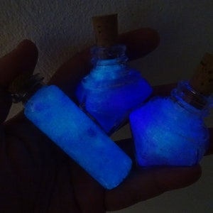 May include: Three small glass bottles with cork stoppers filled with a glowing blue liquid. The bottles are held in a hand.