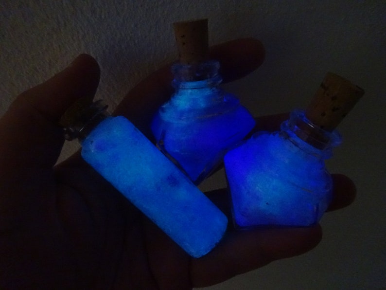 May include: Three small glass bottles with cork stoppers filled with a glowing blue liquid. The bottles are held in a hand.