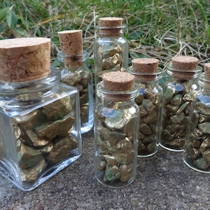 Könnte beinhalten: Sechs Glasbehälter mit Korkverschlüssen, gefüllt mit kleinen, goldfarbenen Steinen. Die Behälter sind in einer Reihe auf einer grauen Oberfläche angeordnet.