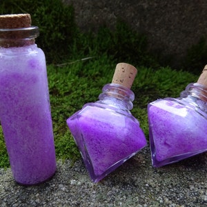 May include: Three glass bottles with cork stoppers filled with a purple and white glittery liquid. The bottles are on a mossy green surface.